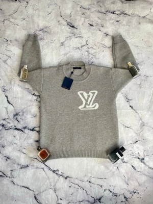 Grå stickad tröja  - Snygg grå stickad tröja med stort vitt logo framtill. Tröjan har rund halsringning och ribbstickade muddar vid ärmslut och nederkant. Perfekt för dig som gillar exklusiva och trendiga plagg.
