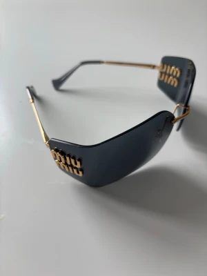 Miu svarta solglasögon med guld - Snygga solglasögon inspirerade med svarta, kantiga glas och tunna guldfärgade skalmar. Glasögonen har en unik design med Miu -logga i guld på sidorna. Perfekt accessoar för en trendig look. 