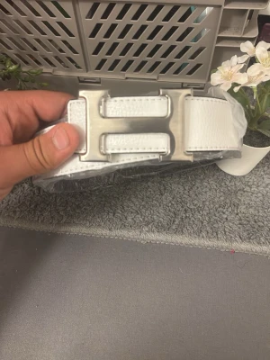 Vitt skärp med H-spänne i silver - Snyggt vitt bälte med ett stort H-format spänne i silverfärgad metall. Bältet är gjort i skinn och har en stilren design som passar till många olika outfits. Perfekt accessoar för att lyfta din look.
