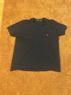 Svart t-shirt från Ralph Lauren - Klassisk svart t-shirt från Ralph Lauren med röd broderad logga på bröstet. T-shirten har rund halsringning och korta ärmar. Perfekt basplagg med stilren design och mjukt bomullsmaterial.