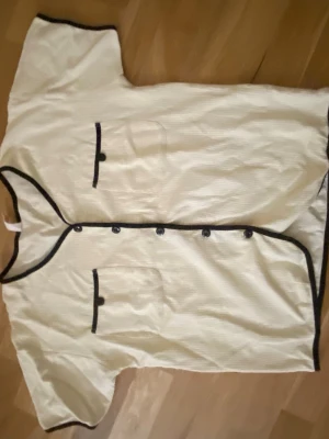 Vit blus med svarta detaljer - Stilren vit blus med svarta kanter och knappar framtill. Två dekorativa fickor på bröstet med svarta detaljer. Kortärmad modell med rak passform, perfekt för en trendig och avslappnad look. Står S i annonsen men den är stretchig och luftig så passar M med garanti! 