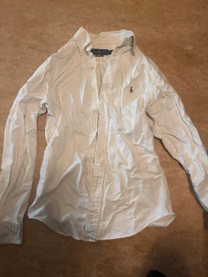 Vit slim fit skjorta från Ralph Lauren - Klassisk vit skjorta från Ralph Lauren i slim fit-modell. Skjortan har långa ärmar, knappar framtill och den ikoniska broderade loggan på bröstet. Tillverkad i mjuk bomull och passar perfekt till en stilren look.