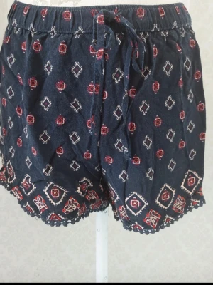 Mönstrade blå shorts med snörning - Snygga mörkblå shorts med röd och vit geometrisk mönster, elastisk midja och snörning framtill. Shortsen har dekorativ kant nertill och är perfekta för sommarens alla äventyr.