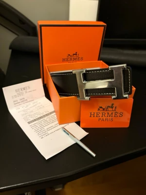Svart H skärp med silver H-spänne - Exklusivt svart skärp från h med ikoniskt H-spänne i silverfärgad metall. Skärpet har vita kontrastsömmar och levereras i original orange H-box med tillhörande certifikat. Perfekt accessoar för att lyfta din outfit.