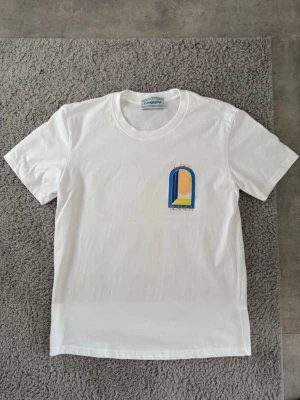Vit t-shirt från Casablanca - Vit t-shirt från Casablanca med färgglatt tryck i blått, gult och orange på bröstet och ryggen. Trycket föreställer ett fönster med texten 'Casa Blanca'. Klassisk passform och rund hals. Perfekt för en stilren och modern look.