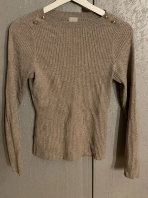 Beige ribbstickad tröja med knappar - Snygg beige ribbstickad tröja med långa ärmar och dekorativa guldfärgade knappar på axlarna. Tröjan har en rund halsringning och är figurnära i modellen. Perfekt för lager-på-lager och enkel att matcha med jeans eller kjol. Passar nån som bär xs