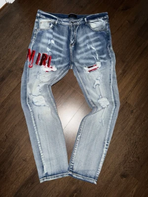 Ljusblå ripped jeans från Amiri - Säljer ett par ljusblå jeans från Amiri med slitningar, hål och röda broderade detaljer. Jeansen har en relaxed fit, klassisk femficksmodell och coola destroyed-effekter framtill. Perfekta för dig som gillar streetwear och statement-plagg.