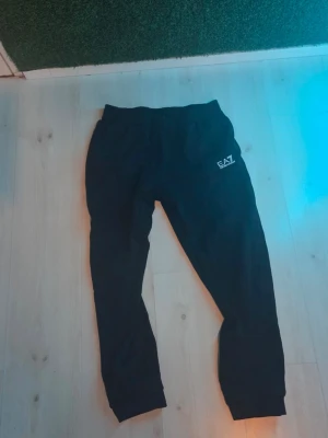 Svarta EA7 mjukisbyxor - Svarta mjukisbyxor från EA7 Emporio Armani med elastisk midja och ribbade benslut. Byxorna har en stilren logga på ena benet och är tillverkade i mjukt material som passar perfekt för en avslappnad look.