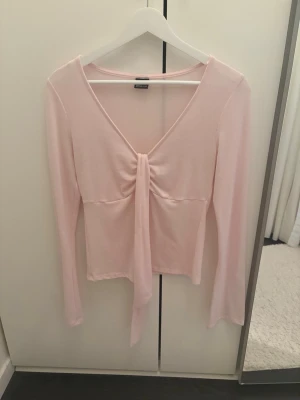 Ljusrosa långärmad topp med knyt - Supersöt ljusrosa topp med långa ärmar och v-ringning. Toppen har en snygg knyt-detalj framtill som ger en feminin touch. Perfekt att styla med jeans eller kjol för en trendig look.