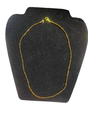 Guldfärgat pärlhalsband - Ett tunt halsband med små guldfärgade pärlor och enkel karbinhake. Halsbandet har en minimalistisk och stilren design som passar perfekt till olika outfits. Materialet är troligen glas eller plastpärlor med metallspänne.