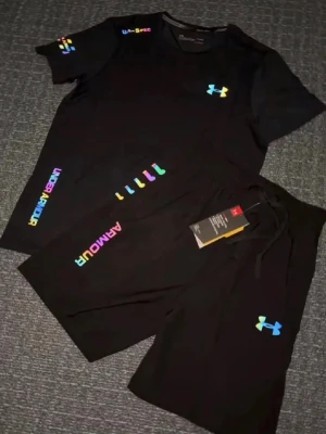 Svart träningsset från Under Armour - Snyggt svart träningsset från Under Armour med t-shirt och shorts. Setet har färgglada reflektiva loggor och textdetaljer i regnbågsfärger. Perfekt för gymmet eller löprundan. Materialet är lätt och andas, vilket gör det bekvämt under träning. Storlek M, helt ny.