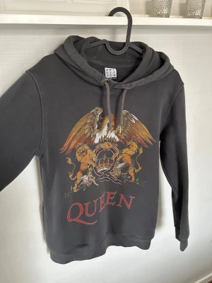 Queen hoodie med tryck - Snygg mörkgrå hoodie med stort Queen-tryck på bröstet. Hoodien har huva med dragsko och fickor på sidan 