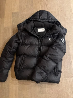 Svart pufferjacka från Calvin Klein - Snygg svart pufferjacka från Calvin Klein med huva och dragkedja. Jackan har två stora fickor med dragkedja och CK-logga på bröstet och ärmen. Perfekt för kalla dagar och riktigt trendig stil.