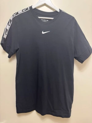 Svart Nike t-shirt med logga - Svart t-shirt från Nike med vit logga på bröstet och vita Nike-band längs axlarna. Klassisk rund hals och korta ärmar. Tillverkad i mjuk bomull för skön känsla och snygg passform.