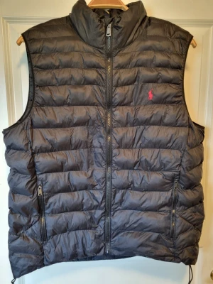 Ralph Lauren väst - Snygg svart dunväst från Polo Ralph Lauren med quiltad design och hög krage. Västen har två dragkedjeförsedda fickor och en diskret röd logotyp på bröstet. Perfekt för lager-på-lager och håller dig varm under kyliga dagar.