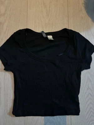 Svart croppad ribbad topp - En svart croppad topp med ribbad struktur och rund halsringning. Toppen är kortärmad och har en tight passform som passar perfekt till jeans eller kjol.