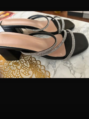Svarta pumps med strass och blockklack - Stilrena svarta pumps med fyrkantig tå, bred blockklack och tunna remmar dekorerade med glittrande strass. Skorna har en öppen design och ljus innersula för extra komfort. Perfekta för att lyfta din outfit med lite extra bling.