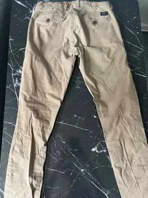 Beiga chinos från selected homme  - Snygga beiga chinos från selected homme med regular fitt passform. Byxorna har två bakfickor med knappstängning och bälteshällor i midjan. Perfect för dig som vill vara finklädd.