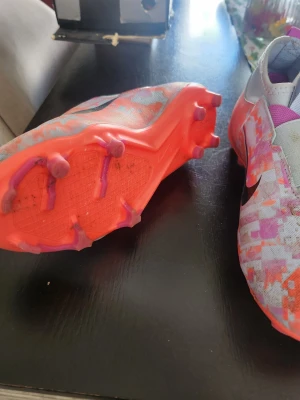 Nike färgglada fotbollsskor - Säljer ett par Nike fotbollsskor med digitalt mönster i orange, lila och grått. Skorna har stora svarta Nike-loggor, elastisk snörning och greppig sula med dobbar i neonorange. Ovandelen är i syntetmaterial och designen är modern och sportig.