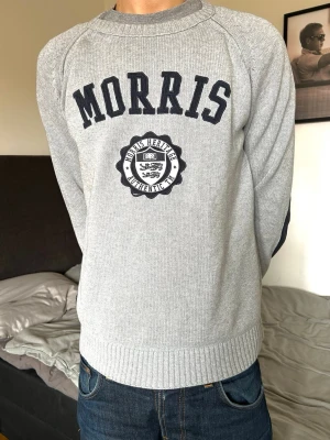 Grå sweatshirt från Morris - Grå sweatshirt från Morris med rund halsringning och svart broderad logga samt emblem på bröstet. Tröjan har långa ärmar och är tillverkad i mjukt bomullsmaterial. Perfekt för en avslappnad och stilren look.