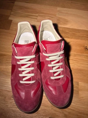 Röda sneakers från Maison Margiela - Säljer ett par röda sneakers från Maison Margiela med en mix av mocka och läder. Skorna har vita snören, beige insida och klassisk gummisula. Snygg retrostil med låg profil och coola detaljer i materialmixen. Ingen låda dustbags 