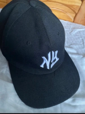 Svart NY keps - Säljer en svart keps med böjd skärm och vit NY-logga broderad framtill. Klassisk modell med ventilationshål och justerbar passform. Perfekt för dig som gillar streetwear och sportig stil.