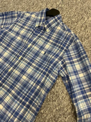 Blårutig skjorta från Polo Ralph Lauren - Klassisk blårutig skjorta från Polo Ralph Lauren med button-down krage och broderad logga på bröstet. Skjortan har långa ärmar, vita knappar och är tillverkad i mjuk bomull. Perfekt för en avslappnad men ändå stilren look.