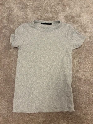 Ljusgrå ribbad t-shirt från lager 157 - En enkel och stilren ljusgrå t-shirt från H&M med ribbad struktur. T-shirten har korta ärmar och rund halsringning, perfekt för en avslappnad look. Materialet känns mjukt och följsamt mot huden.