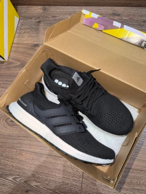 Adidas Ultraboost svarta sneakers - Svarta Adidas Ultraboost sneakers med stickad ovandel och klassiska tre ränder på sidan. Skorna har en vit Boost-sula för extra komfort och svart yttersula. Perfekta för dig som gillar sportig och stilren look.