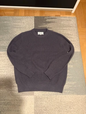 Maison Margiela Hoodie - Säljer en stilren mörkgrå stickad tröja från Maison Margiela. Tröjan har rund halsringning, ribbstickade muddar och är tillverkad i ett mjukt, tjockt material som passar perfekt för kyligare dagar. Enkel och tidlös design som funkar till det mesta.