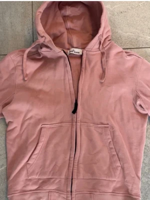 Rosa hoodie Stone Island - Säljer en rosa hoodie från Stone Island med dragkedja framtill och känguruficka. Hoodien har korta ärmar, justerbar huva med snören och är gjord i mjukt bomullsmaterial. Perfekt för en avslappnad streetwear-look.