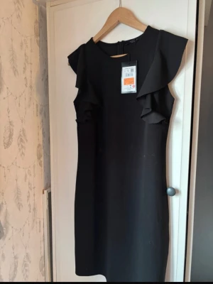 Svart klänning - Stilren svart klänning  med volangdetaljer vid axlarna och ärmlös design. Klänningen har rund halsringning och dragkedja i ryggen. Perfekt för dig som gillar en enkel men elegant look.