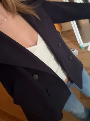 Mörkblå skepparkavaj med stora slag - Snygg mörkblå skepparkavaj med dubbelknäppning och stora slag. Jackan har två framfickor och dekorativa knappar vid ärmsluten. Klassisk och tidlös modell som passar till jeans eller kjol. Materialet ser ut att vara ullblandning.