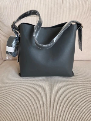 Svart bucket bag i konstläder - Stilren svart bucket bag i slätt konstläder med bred axelrem och magnetlås. Väskan har en innerficka med dragkedja och kommer med extra axelrem. Perfekt för dig som gillar minimalistisk design och vill ha plats för allt du behöver.