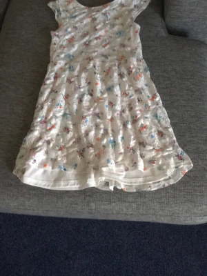 Vit blommig sommarklänning  - Söt vit klänning med färgglatt blommönster i blått, rosa, orange och lila. Klänningen har rund halsringning och är ärmlös, perfekt för varma dagar. Tillverkad i ett lätt och luftigt material som känns bekvämt mot huden. Det finns även en dragkedja på baksidan!