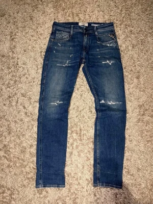 Blå slitna jeans från Replay - Säljer ett par blå jeans från Replay med snygga slitningar och tvättade detaljer. Klassisk femficksmodell med raka ben och låg midja. Jeansen har en cool vintage-look och är tillverkade i mjuk denim för skön känsla.