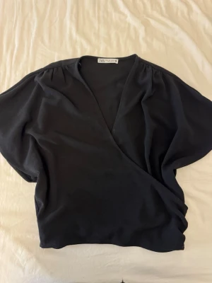 Svart omlottopp från Zara - Snygg svart omlottopp från Zara med korta ärmar och v-ringad hals. Toppen har en avslappnad passform och är tillverkad i mjukt material som känns skönt mot huden. Perfekt att styla med jeans eller kjol för en trendig look.