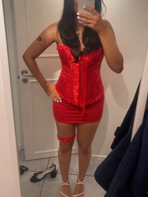 Röd korsett med spetsdetaljer - Snygg röd korsett med satinkänsla och dekorativa spetsdetaljer. Framtill finns det metall knappar man knäpper och baktill är det snörning samt volangkant upptill och nedtill. Korsetten har ett vackert mönster i tyget och ger en feminin siluett. Perfekt för dig som vill sticka ut. Korsetten har en figurnära passform står stl L men skulle säga att den motsvarar Xs-S.