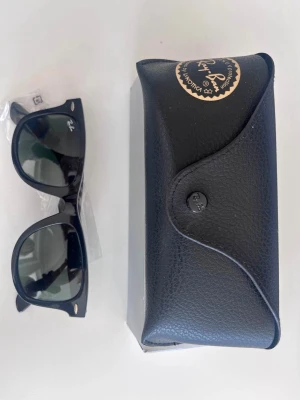 Svarta Ray-Ban solglasögon - Klassiska svarta Ray-Ban Wayfarer solglasögon med ikonisk fyrkantig form och mörka glas. Säljs med original svart fodral i skinnimitation. Perfekta för dig som vill ha en tidlös och stilren look.