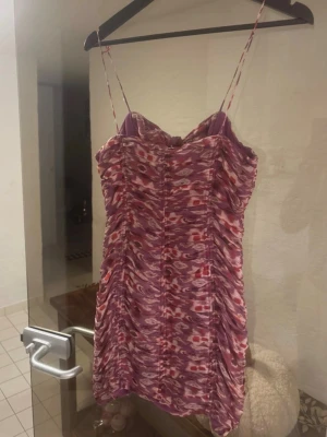 Mini dress - Kort klänning med smala axelband i lila och rosa toner. Klänningen har ett abstrakt mönster, rynkade detaljer och en rosett baktill med en liten cut-out. Materialet känns lätt och luftigt, perfekt för varma dagar.