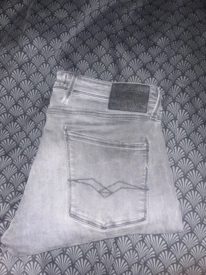 Ljusgrå slim fit jeans från Replay - Säljer ett par ljusgrå slim fit jeans från Replay med klassisk femficksdesign och snygg liten wash. Jeansen har en diskret logga på bakfickan och ett svart läderpatch i midjan. Perfekta för dig som gillar en clean och modern look. Jeansen är i hyperflex. Längd 30 midja33.