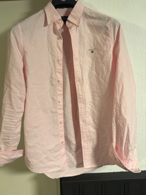 Ljusrosa skjorta från Gant - Klassisk ljusrosa skjorta från Gant med button-down krage och broderad logga på bröstet. Skjortan har långa ärmar, vita knappar och är tillverkad i mjuk bomull. Perfekt för en stilren och avslappnad look.