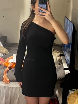 Svart oneshoulder kortklänning - Snygg svart oneshoulder klänning med lång ärm på ena sidan och axelbandslös på andra. Kort modell med figurnära passform, perfekt för dig som gillar stilrena och trendiga plagg. Materialet känns mjukt och följsamt.