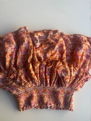 Mönstrad kort volangkjol i orange och rosa. - Väldigt snyggt inför sommaren! Kort volangkjol med smock i midjan och ett färgstarkt mönster i orange, rosa och inslag av gult. Kjolen har flera lager volanger och är gjord i ett lätt och luftigt material, perfekt för sommarens varma dagar. 