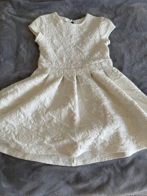 Vit kortklänning med blommönster - Supersöt vit kortklänning med blommigt strukturmönster och rund hals. Klänningen har korta ärmar och en lätt utställd kjol som ger en fin siluett. Perfekt för sommarens alla tillfällen!