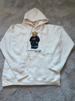 Vit hoodie Polo Bear Ralph Lauren - Vit hoodie från Ralph Lauren med den ikoniska Polo Bear-trycket framtill. Hoodien har dragsko i huvan och en stor magficka. Tillverkad i mjukt bomullsmaterial som är skönt att bära. Perfekt för dig som gillar klassisk streetwear med en twist. Ny utan prislapp