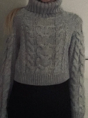 Grå kabelstickad polotröja - Mysig grå kabelstickad polotröja med croppad passform och breda ärmar. Tröjan har en hög polokrage och grova flätmönster både fram och på ärmarna. Perfekt för kalla dagar och enkel att styla till olika outfits.