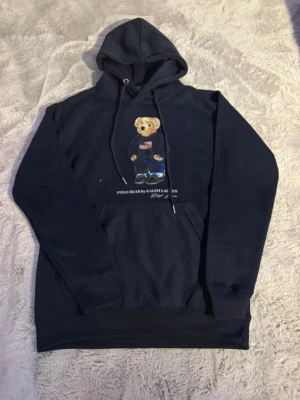 Svart Polo Bear hoodie från Ralph Lauren - Säljer en svart hoodie från Ralph Lauren med den ikoniska Polo Bear-trycket på bröstet. Hoodien har huva med dragsko och en stor magficka. Materialet är mjukt och skönt, perfekt för en avslappnad stil. Klassisk och tidlös design som alltid funkar. Ny utan prislapp. Dock har öljetter lossnat lite på ena sidan