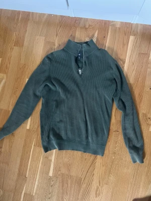 Olivgrön stickad halvzip-tröja - Säljer en olivgrön stickad tröja med halv dragkedja och hög krage. Tröjan har lång ärm och ribbade muddar vid ärmslut och nederkant. Perfekt för lager på lager och chill stil.