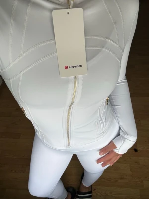 Vitt träningsset från Lululemon - Snyggt vitt träningsset från Lululemon med jacka och tights.😍Jackan har dragkedja framtill och guldfärgade detaljer vid fickorna. 🤍Setet är gjort i ett stretchigt och mjukt material som passar perfekt till gymmet eller yoga, ÄLSKAR verkligen detta settet😍.                                                                    ‼️OBS‼️- Jag har i både storlek 6 & 4, storlek 4 = xxs/xs & storlek 6 = xs/s 